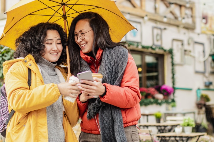 two women wearing rain jackets under yellow umbrella looking at phone