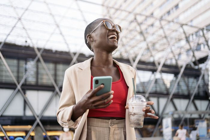 Young Black woman with shaved head in sunglasses and a cream blazer holding a cellphone in turquoise case and a frappuccino looking up and to the left